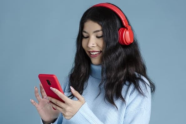 A woman with red headphones and a blue sweater smiles at her smartphone while scheduling posts to Instagram.